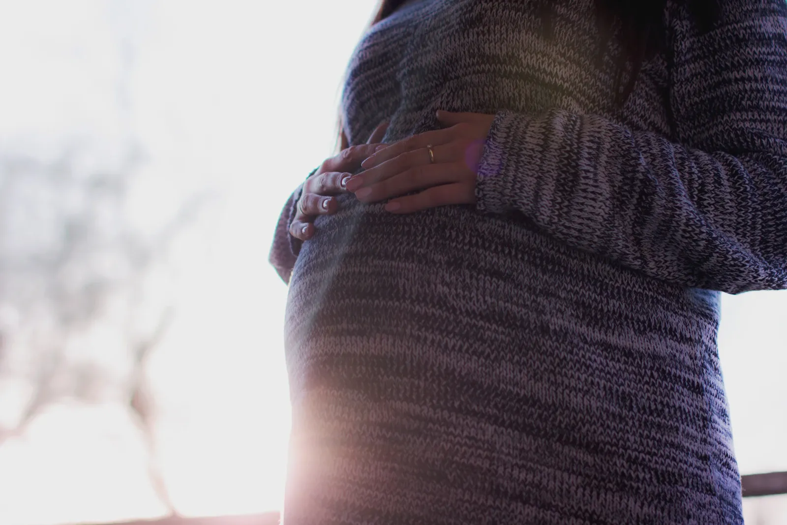 Schwangere im Strickpullover hält die Hände im warmen Gegenlicht auf dem Bauch